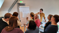 Das Bild zeigt eine Gruppe Anwärter*innen des Studienseminars für BBS Neuwied in einem Raum beim Umsetzen der Übung Collaboration-Puzzle. Dabei stehen sie vor einem Flipchart.