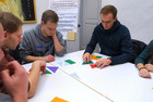 Bei der Übung „Teamquadrat“ werden aus farbigen Puzzleteilen zehn gleich große Quadrate auf einem Tisch gelegt. Auf dem Bild legen 3 Anwärter sehr konzentriert die Quadrate, links sieht man teilweise einen weiteren Anwärter, rechts teilweise noch eine Anwärterin.