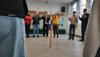 Mit Hilfe von Schnüren und einem Kran wird in einem Seminarraum ein Holzklotz auf einen Turm mit 4 Holzklötzen gestellt. Im Hintergrund sieht man 6 Auszubildende in Aktion.